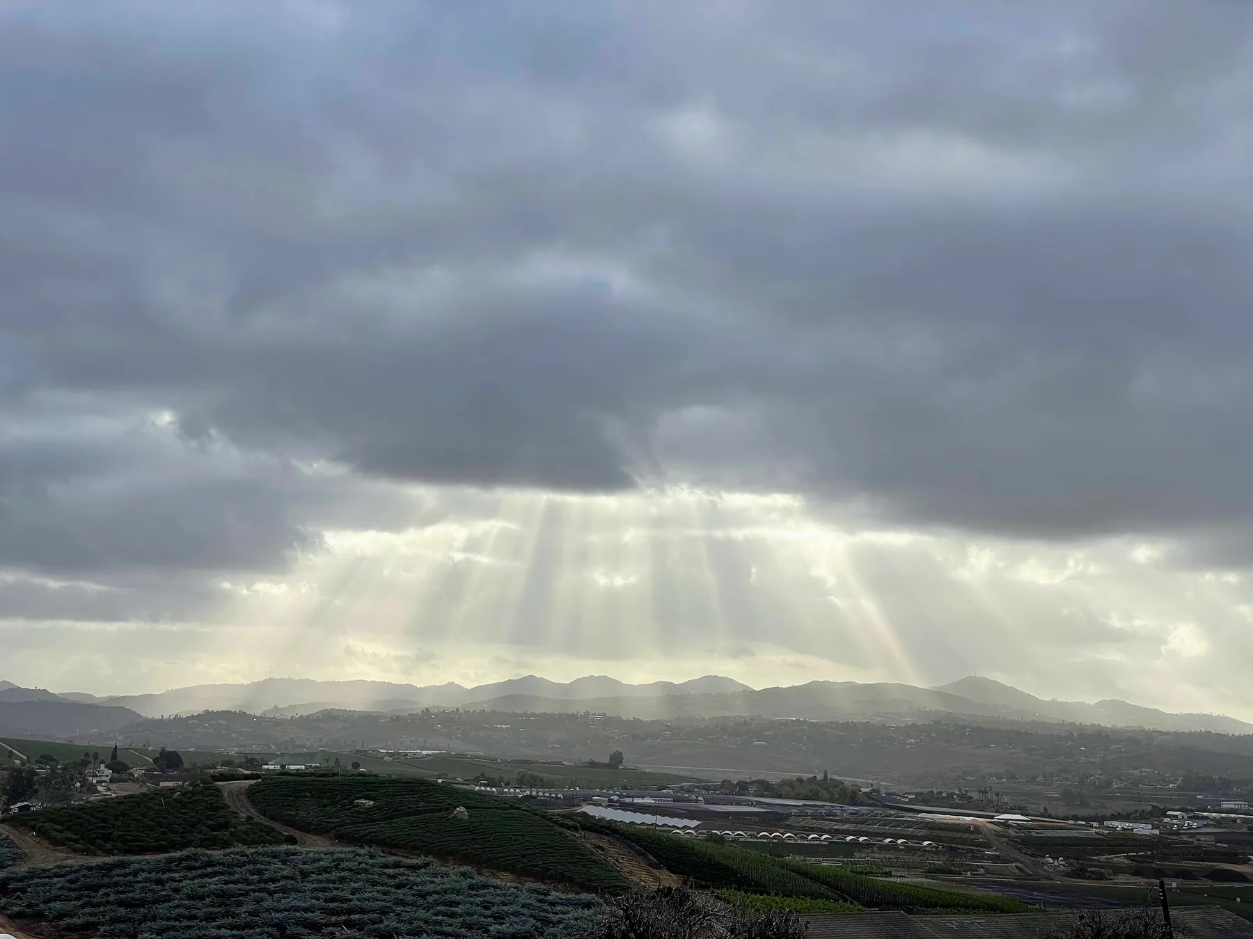 Rows of crops and greenhouses across South Morro Hills, with sunbeams breaking through clouds over distant hills